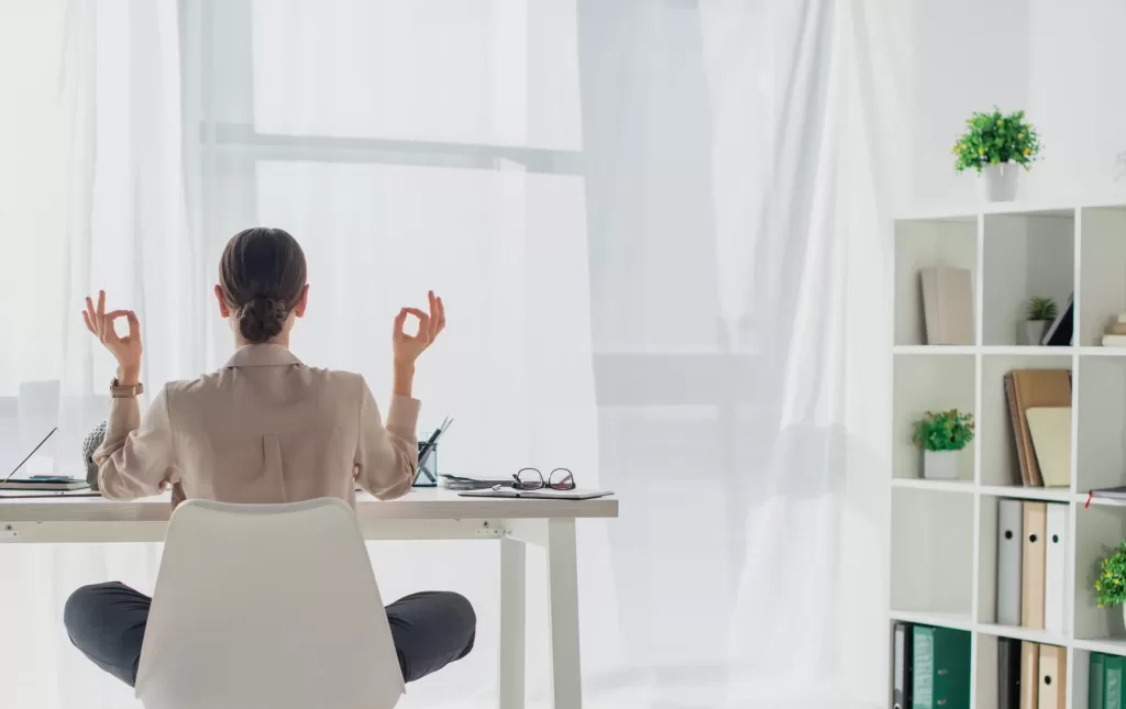 Back view of businesswoman meditating in lotus pose with gyan mudra at workplace