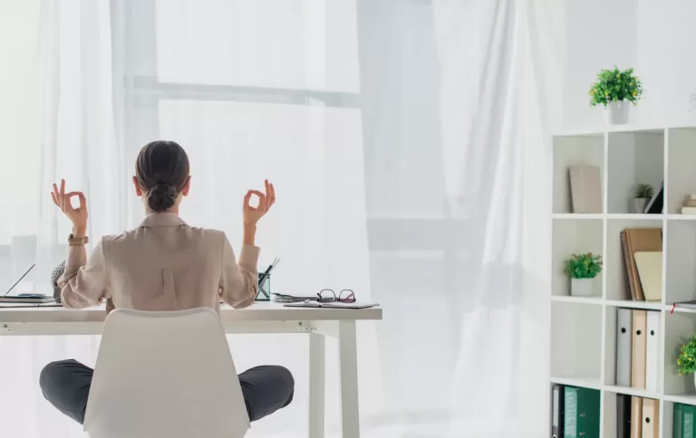 Back view of businesswoman meditating in lotus pose with gyan mudra at workplace
